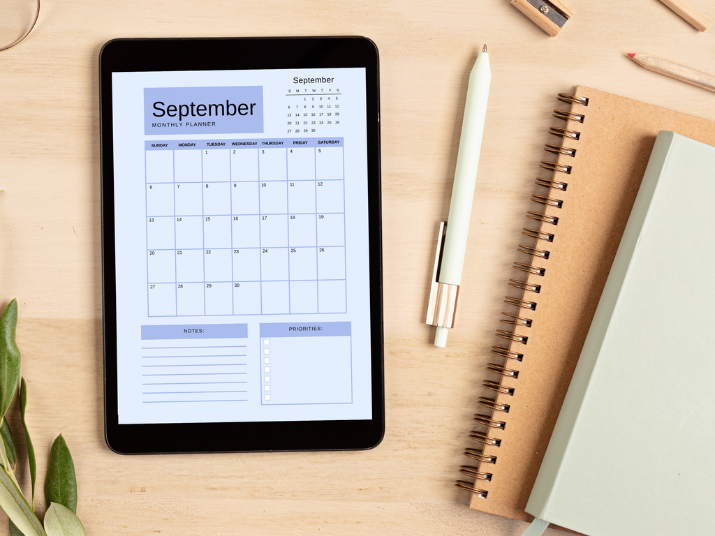 Digital tablet displaying a monthly planner on a wooden surface with a pen and notebook.