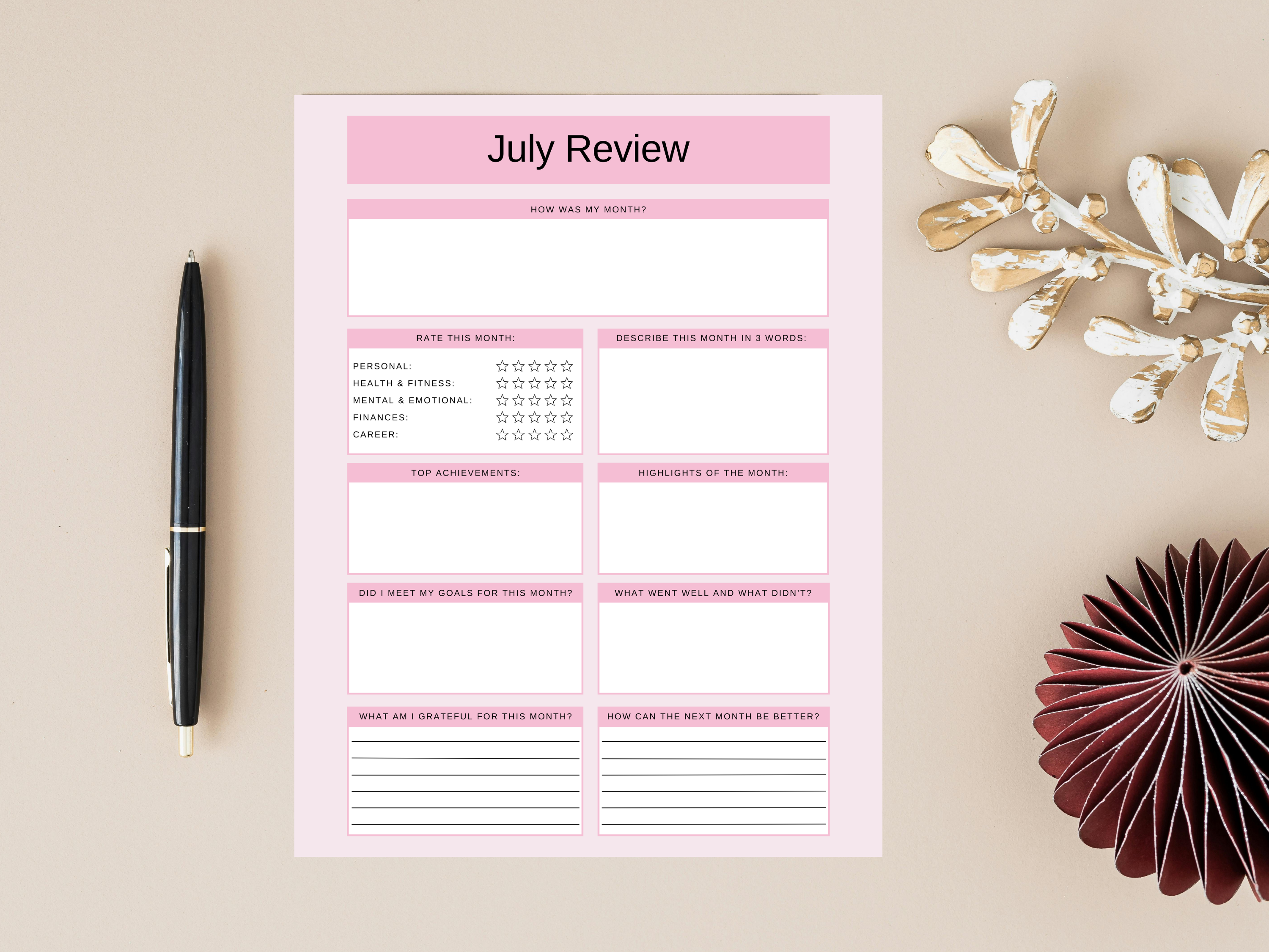 Pink July review planner sheet on a beige surface with a pen and decorative items.